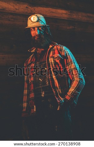 Male Miner Wearing Lit Safety Helmet Lamp with Hand in Pocket Leaning Against Wooden Wall in Plaid Shirt and Looking to the Side