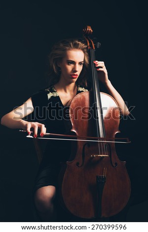 Young woman playing the cello in a classical concert or recital sitting in the darkness with a serious expression as she awaits her cue to start playing