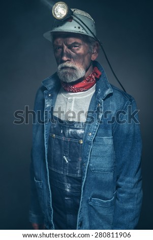 Close up Bearded Senior Male Mine Worker, Wearing Denim Jacket and Helmet with Lamp, Looking Straight at the Camera in a Serious Facial Expression.