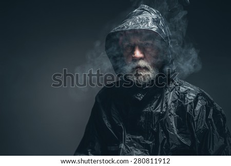 Close up Pensive Old Bearded Guy in Black Rain Jacket with Cigarette Smoke Against Black Background.
