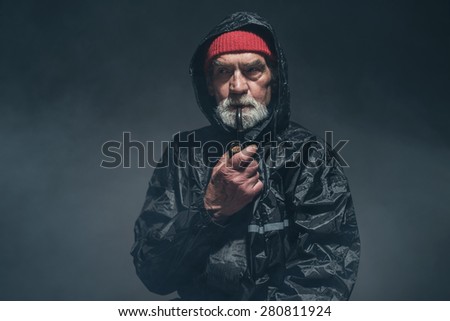 Bearded Elderly Guy in Black Winter Outfit Smoking Using Pipe While Looking Into Distance and Thinking of Something. Captured in a Fuzzy Studio.