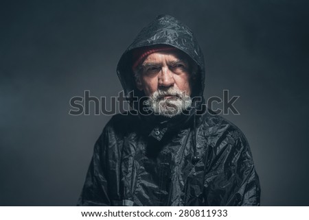 Close up Serious Bearded Old Man in Black Waterproof Coat, Looking Into Distance Against Black Background.