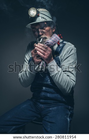 Portrait of a Dirty Adult Gold Miner Sitting on a Stool with his Helmet, Lighting his Cigarette While Looking at the Camera.