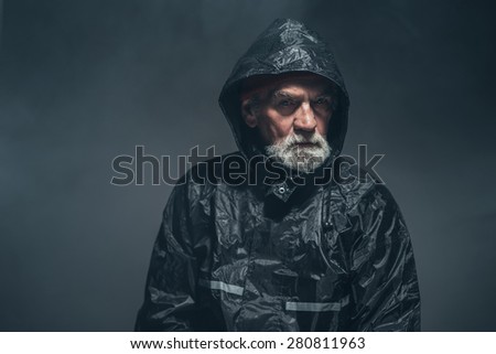 Close up Humorless Bearded Old Man in Black Rain Jacket Looking Straight at Camera Against Black Background.