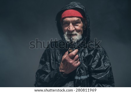 Close up Smiling Bearded Guy in Black Winter Raincoat, Smoking Using a Tobacco Pipe While Looking at the Camera. Captured in Studio with a Fuzzy Black Background.
