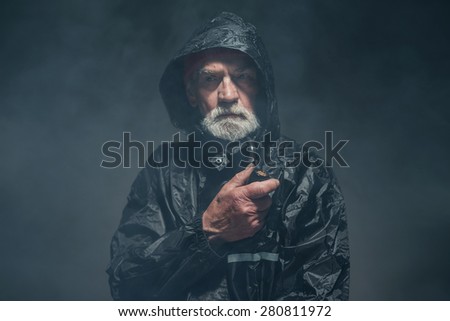 Close up Bearded Old Guy in Black Water-Resistant Jacket, Holding a Smoking Pipe While Looking Into Distance Seriously. Capture in a Fuzzy Studio.