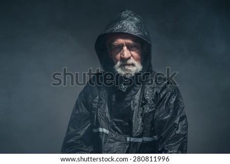 Portrait of a Bearded Senior Man in Black Waterproof Jacket, Looking at the Camera on a Fuzzy Black Background in the Studio.