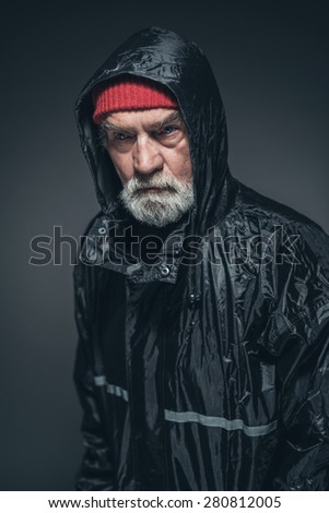 Close up Elderly Man with White Facial Hair, Wearing Red Bonnet and Black Rain Slicker, Looking Straight at the Camera. Captured in Studio with Black Background.