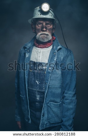 Close up Portrait of a Bearded Senior Male Miner in Denim Jacket and Helmet with Dirty Face, Looking at the Camera Seriously Inside a Fuzzy Studio.