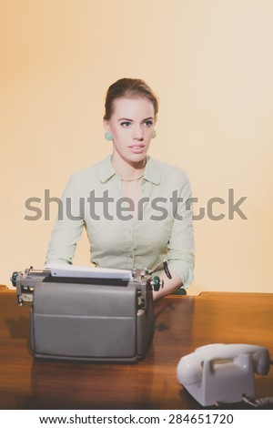 Distracted retro 1950 blonde secretary woman sitting behind desk working on typewriter. High angle view.