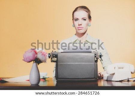 Serious vintage 1950 blonde secretary woman sitting behind desk working on typewriter.
