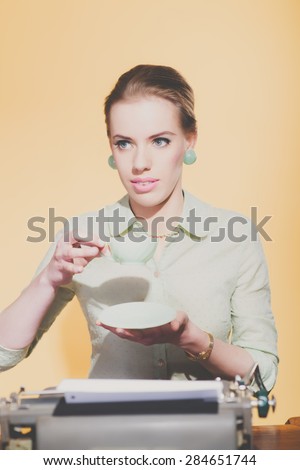 Retro 50s young blonde secretary woman sitting behind desk drinking cup of tea.