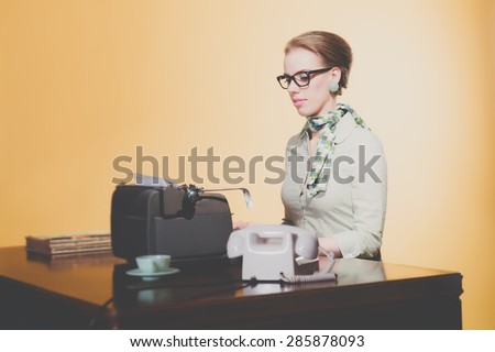 Vintage 1950 young secretary woman behind desk typing on typewriter wearing black glasses.