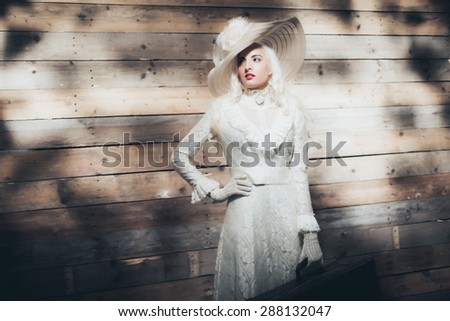 Elegant blond lady in stylish vintage fashion wearing a hat gloves and long gown standing in dappled sunlight in front of a wooden wall with her hand holding a leather suitcase