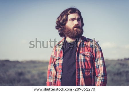 Handsome bearded man wearing a red plaid shirt standing outdoors in nature looking off to the right of the frame with a thoughtful expression