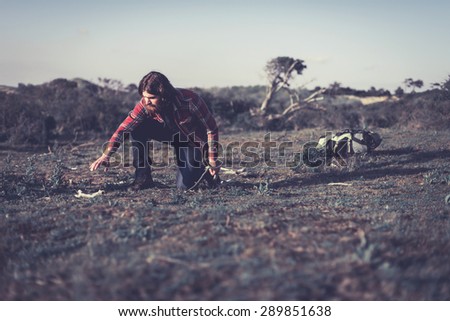 Hiker collecting firewood to make a campfire to cook a meal leaning down to pick up dry branches with his backpack on the ground behind him
