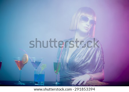 Glamorous and Sophisticated Woman with Red Hair Wearing Shiny Retro Gown and Sunglasses Leaning on Bar Covered in Cocktails in Smoky Disco Night Club Bar