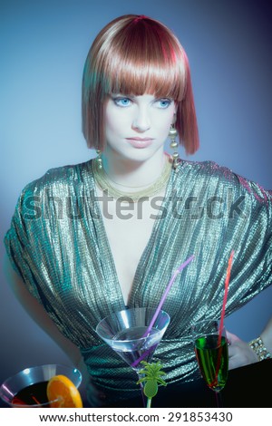 Glamorous Woman with Red Hair Wearing Shiny Retro Gown and Standing at Bar Covered with Drink Cocktails in Disco Night Club