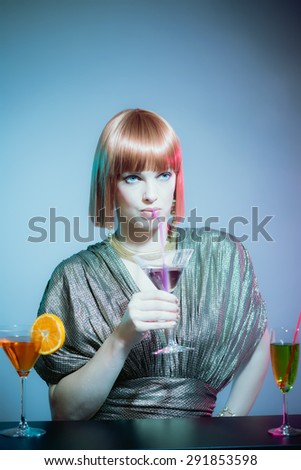 Glamorous Woman with Red Hair Wearing Shiny Retro Gown and Sipping Cocktail Drink from Straw, Standing at Bar in Disco Night Club