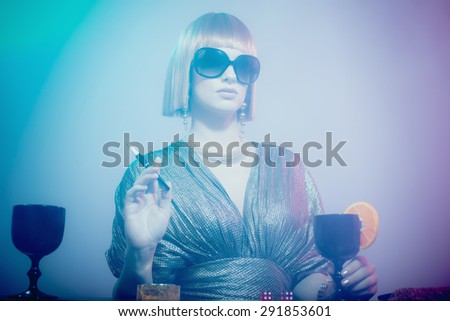 Glamorous and Sophisticated Woman with Red Hair Wearing Sunglasses and Shiny Retro Gown Standing at Bar Smoking Cigarette and Gambling in Smoky Disco Night Club