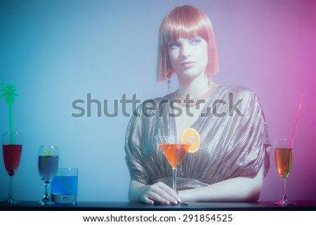 Glamorous and Sophisticated Woman with Red Hair Wearing Shiny Retro Gown with Fruity Martini Drink Leaning on Drink Lined Bar in Disco Club