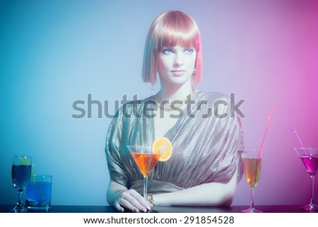 Glamorous and Sophisticated Woman with Red Hair Wearing Shiny Retro Gown with Fruity Martini Drink Leaning on Drink Lined Bar in Disco Club