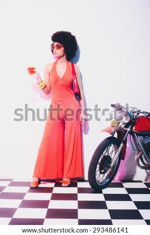 Elegant Young Lady in an Orange Fashion Standing Beside a Motorcycle with a Cocktail on her Hand, Looking Into Distance.