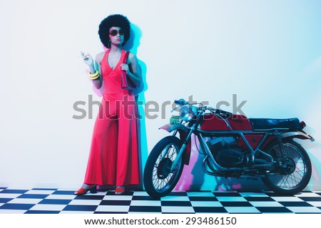 Elegant Young Woman Holding a Cigarette While Posing Beside her Vintage Motorcycle on a White Wall Background.