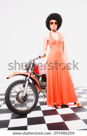 Stylish Young Woman in Orange Dress, Posing on her Vintage Motorcycle on a Checkered Floor Against White Background.