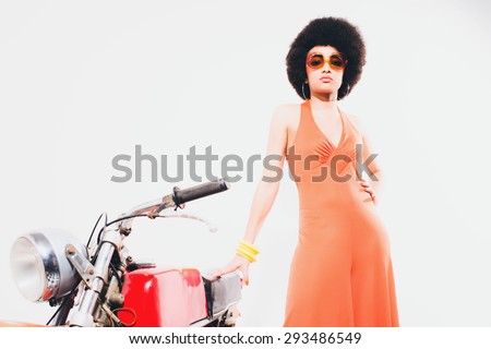 Elegant Young Woman in an Orange Fashion, Standing Beside her Motorcycle and Looking at the Camera Against Off-White Background.