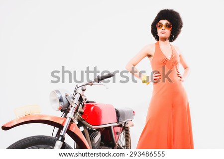 Elegant Young Woman in an Orange Fashion, Standing Beside her Motorcycle and Looking at the Camera Against Off-White Background.