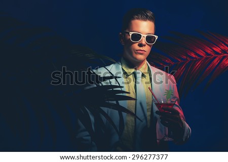 Half Body Shot of a Fashionable Young Man in Business Suit with Sunglasses, Holding a Cocktail Drink Between Palm Leaves Against Dark Blue Gray Background.