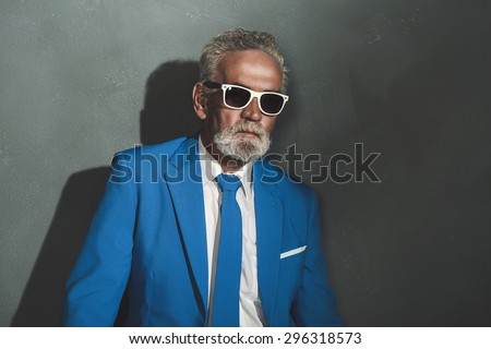 Half Body Shot of a Senior Bearded Businessman Wearing Blue Formal Suit with Sunglasses, Leaning Against Plain Gray Wall.