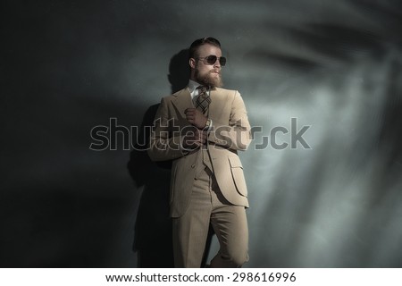 Elegant man adjusting the sleeve of his jacket as he stands in a pair of sunglasses leaning against a grey wall with offset beam of light for copyspace