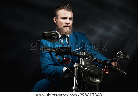Handsome bearded businessman in a blue suit sitting waiting on a motorbike in the darkness looking thoughtfully off to the side, with copyspace