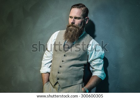 Pensive Bearded Man in Formal Wear Looking Into the Distance with Hands in the Pockets Against Gray Wall.