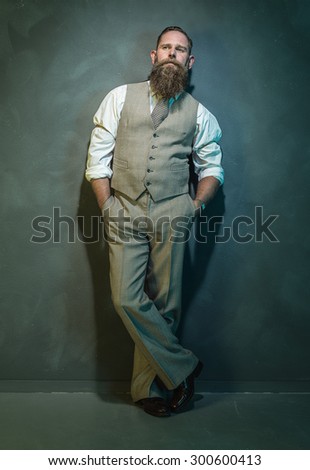 Full Length Shot of a Thoughtful Bearded Man in Formal Outfit, Looking into the Distance While Leaning Against Wall with Hands in the Pockets and Legs Crossed.