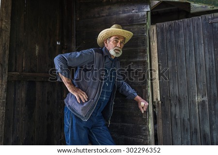 Senior farmer or farmhand in an old rustic wooden barn standing leaning against the wall and door with a serious expression as he waits for someone
