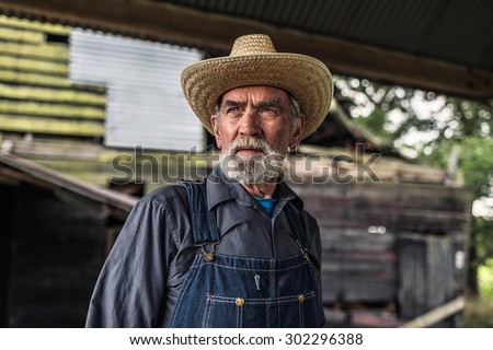 Old farmer standing in front of a rustic dilapidated wooden barn staring thoughtfully off to the side