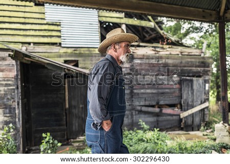 Old farmer standing in front of a rustic dilapidated wooden barn with his hands in his pocket staring thoughtfully off to the side