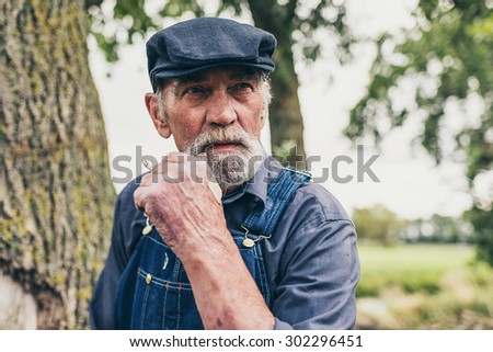 Senior grey-haired bearded country farmer standing smoking in his cloth cap and denim dungarees as he stares thoughtfully into the distance