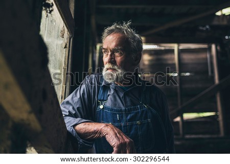 Elderly man staring out of a rustic wooden window in an old rural barn or house with a thoughtful serious expression