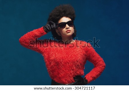 Half Body Shot of a Stylish Young Woman with Afro Hairstyle, Wearing Fuzzy Tops with Sunglasses against Blue Wall Background.
