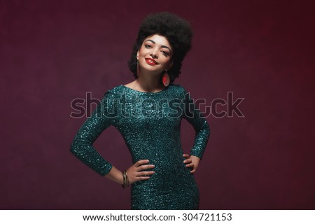 Half body shot of a stylish young woman wearing sparkling green dress, looking at the camera with her both hands on waist. Isolated on purple background.