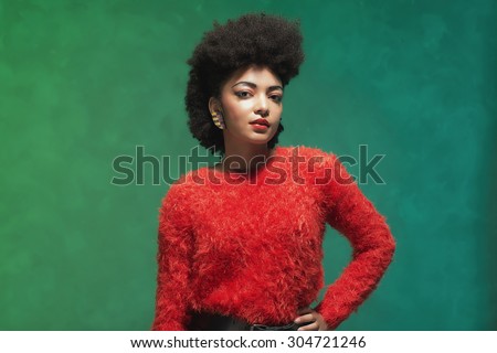 Half body Shot of a Stylish Young Woman with Afro Hair, Wearing Furry Red Shirt and Black Shorts, Looking at the Camera Against Green Wall