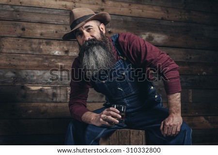 Vintage worker man with long gray beard in jeans dungarees holding whiskey. Sitting on wooden crate in barn.