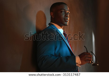 Retro african american businessman in blue suit holding sunglasses. Leaning against gray wall.