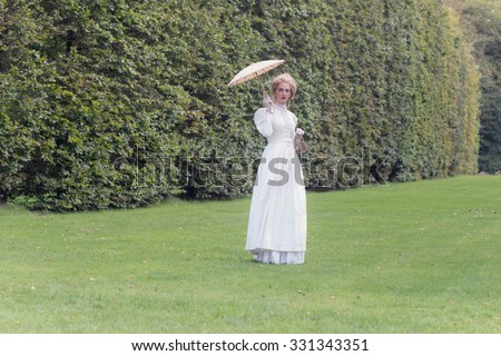 Victorian fashion woman holding parasol standing on lawn with tall hedge.