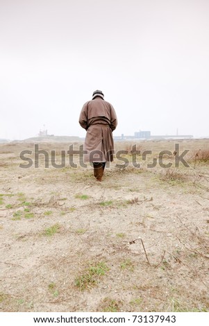 Senior man with raincoat and hat walking on sand with grass. Open space. Lost.
