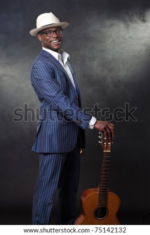 Black jazz musician wearing suit and white hat standing with guitar. Smoking cigarette and wearing glasses.